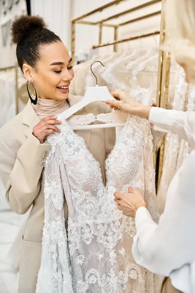 Dos mujeres jóvenes, una hermosa novia y un ayudante de tienda útil, examinando un vestido de novia en una percha en una boutique. - foto de stock