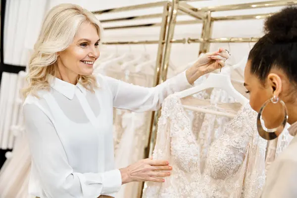 Una joven novia hermosa examinando un vestido en un estante mientras hace compras para el día de su boda con un asistente de tienda. - foto de stock