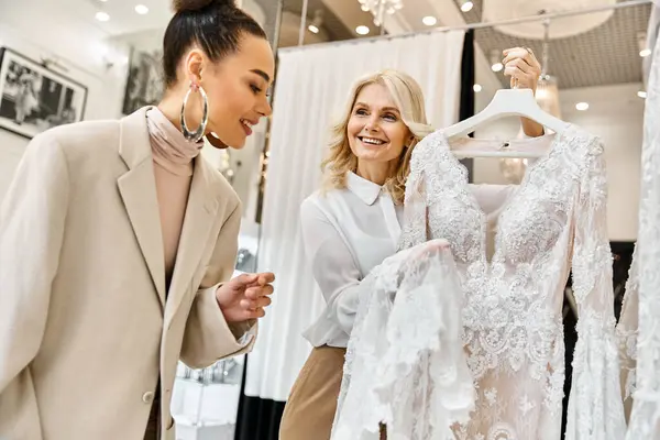 Two women, a young beautiful bride and a shop assistant, browsing elegant dresses in a store. — Stock Photo