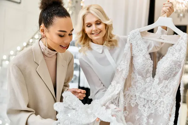 Dos mujeres admirando un vestido blanco en una percha en una tienda de novias. La futura novia y el asistente de la tienda están discutiendo el vestido. - foto de stock