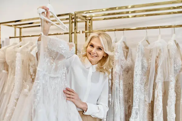 Une belle assistante shopping d'âge moyen se tient gracieusement devant un rack de robes de mariée élégantes dans un salon de mariée. — Photo de stock