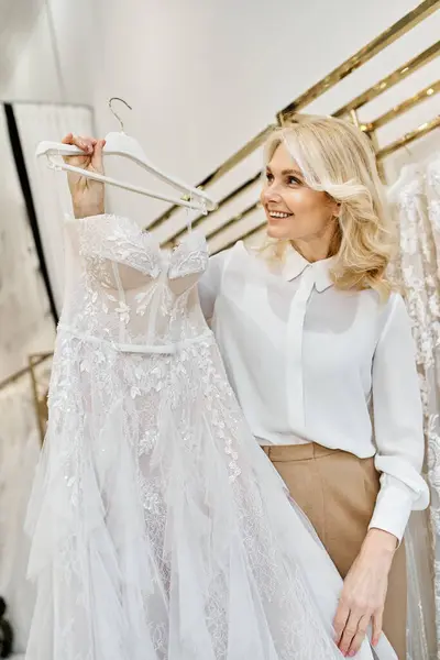 Красивий помічник шопінгу середнього віку у весільному салоні, який тримає ніжну білу сукню з турботою. — стокове фото