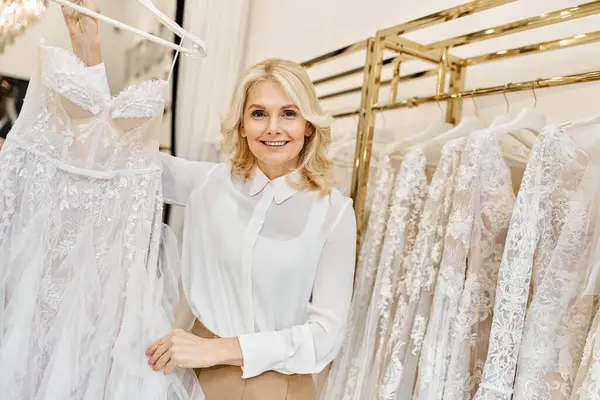 Um lindo assistente de compras de meia-idade está diante de um rack de vestidos em um salão de casamento, ajudando os clientes. — Fotografia de Stock