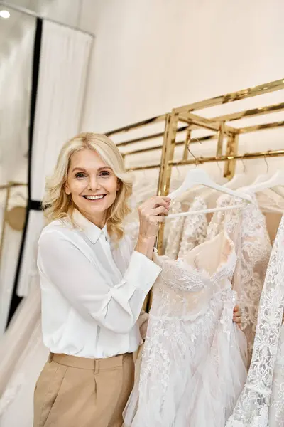Un hermoso asistente de compras de mediana edad se coloca elegantemente delante de un estante de vestidos de novia en un salón de novias. - foto de stock