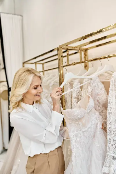 A middle-aged beautiful shopping assistant helps a woman browse through wedding dresses on a rack in a salon. — Stock Photo