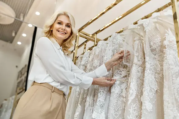 A middle-aged beautiful shopping assistant stands in front of a rack of dresses in a wedding salon, assisting customers. — Stock Photo