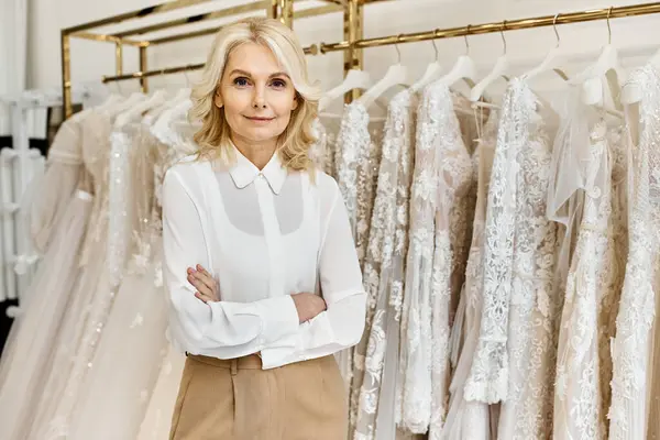 Eine schöne Verkäuferin mittleren Alters steht in einem Brautsalon vor einem Regal mit eleganten Brautkleidern. — Stockfoto
