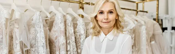 Mediana edad hermosa asistente de compras de pie delante de un estante de vestidos en un salón de bodas. - foto de stock