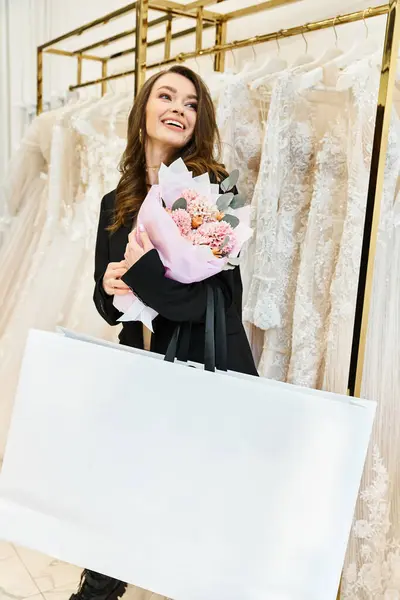 Joven novia morena sosteniendo ramo en frente de rack de vestidos de novia en salón de novia. - foto de stock