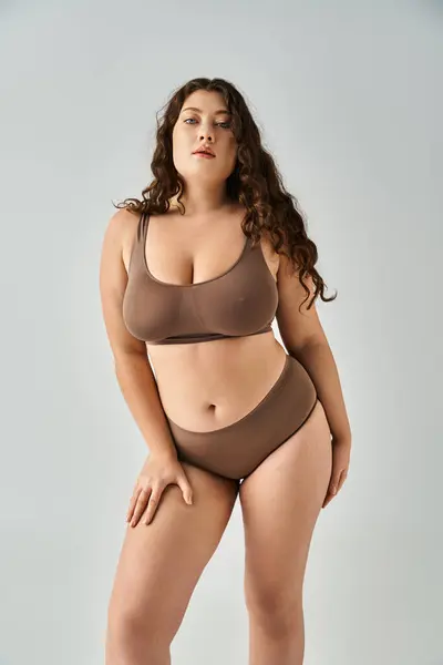 Hermosa mujer con curvas en ropa interior con el pelo castaño inclinado hacia adelante y posando sobre fondo gris - foto de stock