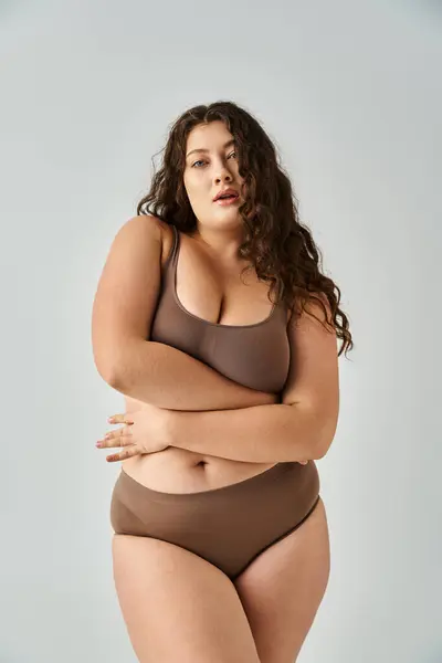Alluring plus size young woman in underwear with curly brown hair hugging herself on grey background — Stock Photo