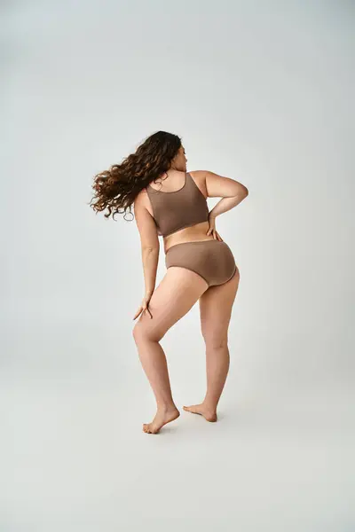 Plus la taille jeune femme en sous-vêtements avec les cheveux bruns bouclés posant du dos sur fond gris — Photo de stock