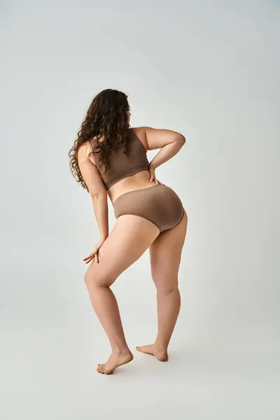Curvy young girl in underwear with curly brown hair posing from back on grey background — Stock Photo