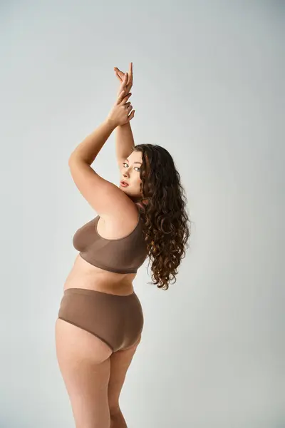 Attractive curvy woman in underwear with curly brown hair posing from back and putting hands to up — Stock Photo