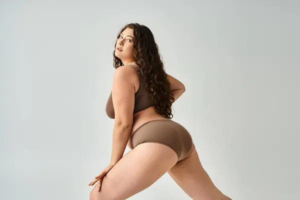 Seductive curvy woman in underwear with curly brown hair posing from sideways on grey background — Stock Photo