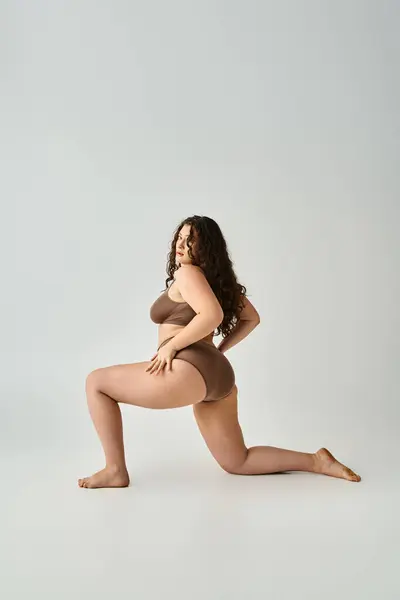 Atractiva mujer de talla grande con el pelo castaño rizado posando de lado con las piernas sobre fondo gris - foto de stock