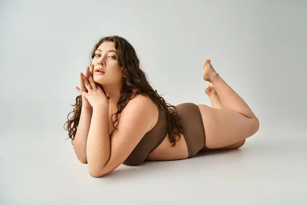 Hermosa chica de talla grande en lencería marrón con pelo rizado seductora tumbado sobre fondo gris - foto de stock