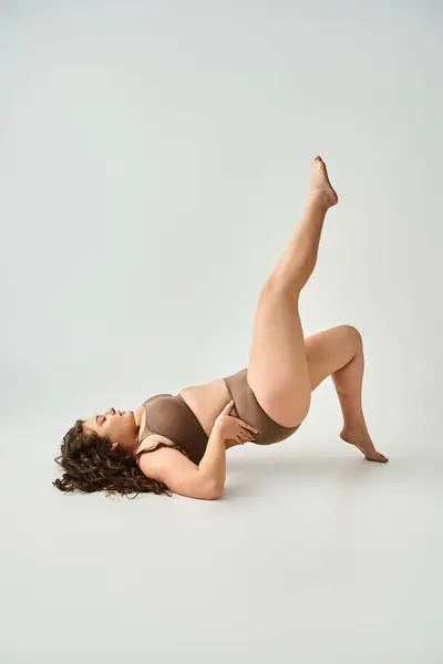 Charming plus size woman in brown lingerie with curly hair posing and putting leg to up — Stock Photo
