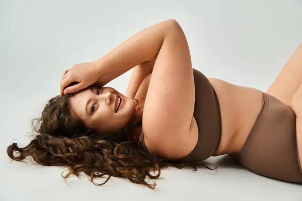 Sorridente giovane donna curvy in lingerie marrone con capelli ricci sdraiati e mettendo mano sulla testa — Foto stock
