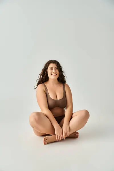 Cheerful curvy young woman in brown underwear sitting cross legged against grey background — Stock Photo