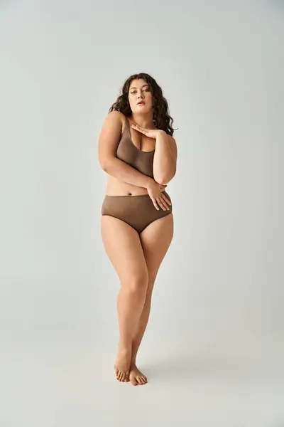 Seductive plus size woman in brown lingerie with curly hair hugging herself on grey background — Stock Photo