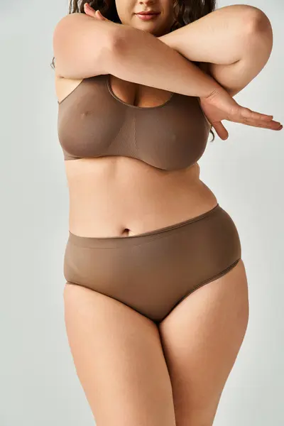 Cropped shot of body plus size young woman in brown lingerie crossing arms to up on grey background — Stock Photo