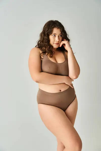 Bonita mujer con curvas en lencería marrón con el pelo rizado posando con los brazos sobre fondo gris - foto de stock