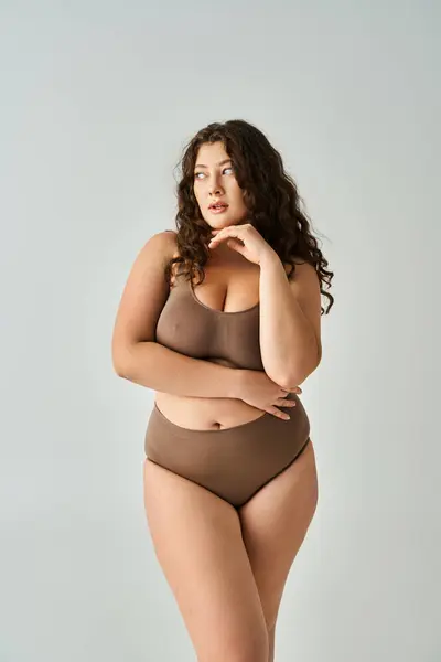 Encantadora mujer con curvas en lencería marrón con el pelo rizado posando con los brazos y mirando a un lado - foto de stock
