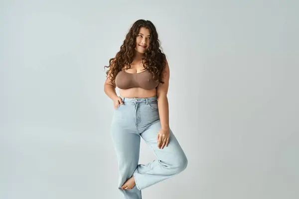 Smiling plus size young woman in brown bra and blue jeans with curly hair posing with bent leg — Stock Photo