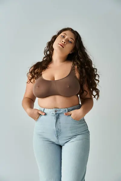 Beautiful curvy woman in brown bra and blue jeans posing with hands in pockets and leaning head — Stock Photo