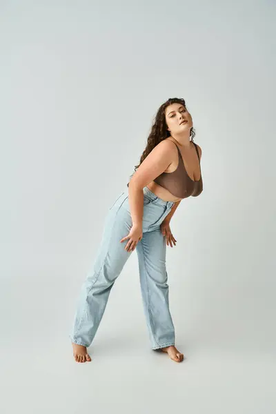 Beautiful plus size woman in brown bra and blue jeans leaning to forward on grey background — Stock Photo
