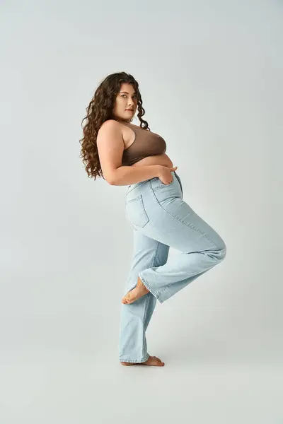 Довольно пышная женщина в коричневом бюстгальтере и синих джинсах склоняется к спине с изогнутой ногой на сером фоне — стоковое фото