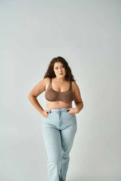 Cheerful plus size woman in brown bra and blue jeans posing with hands in pockets on grey background — Stock Photo