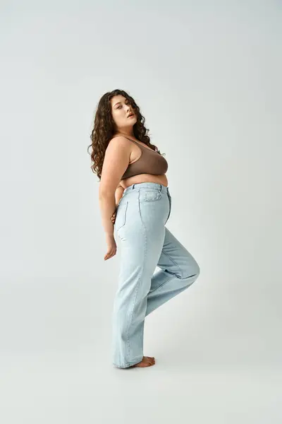 Attraente plus size donna in reggiseno marrone e jeans blu con le mani dietro la schiena posa lateralmente — Foto stock