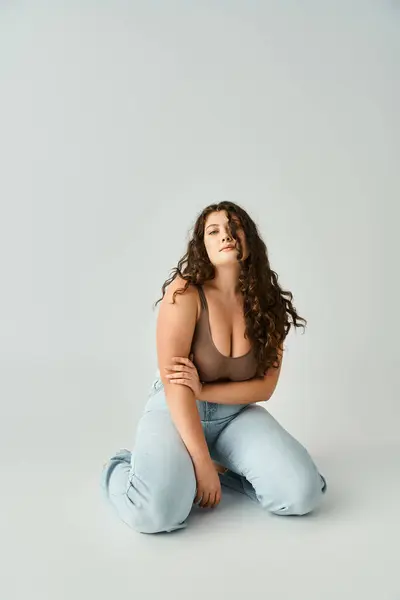 Seducente plus size giovane donna in reggiseno marrone e jeans blu seduta e toccante con mano alle braccia — Foto stock