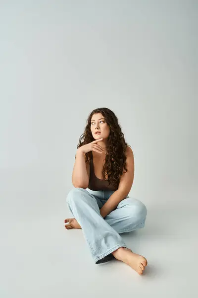 Attractive plus size young girl in brown bra and blue jeans sitting and touching finger to chin — Stock Photo