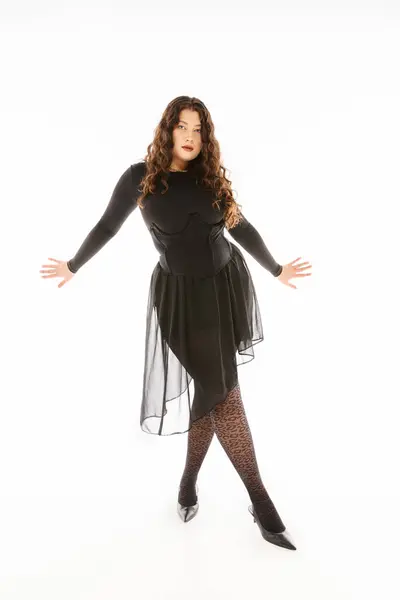 Mujer joven con curvas glamurosas en traje elegante negro con las manos en los lados y las piernas cruzadas en gris — Stock Photo