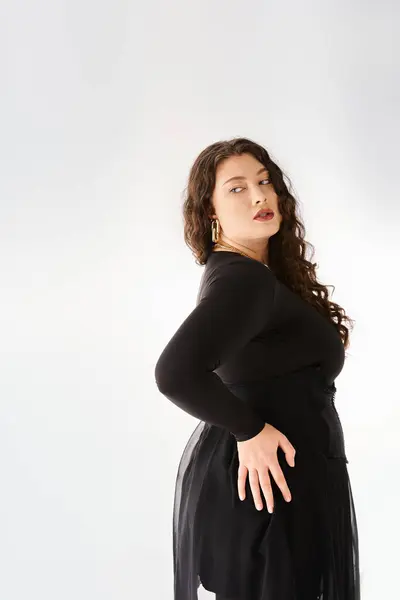 Séduisante femme plus taille en tenue élégante noire avec des cheveux bouclés posant du dos avec la main sur la hanche — Photo de stock
