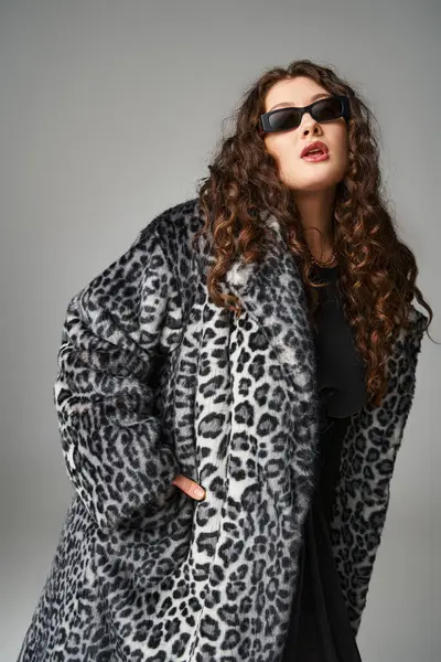 Mujer joven elegante más tamaño en abrigo de piel de leopardo y gafas de sol posando sobre fondo gris - foto de stock