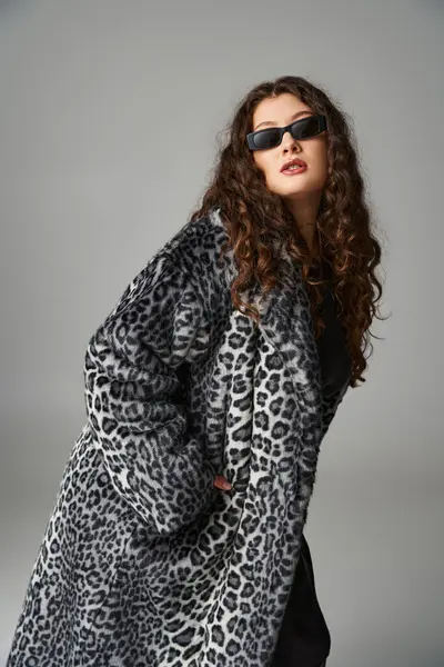 Glamour plus taille femme en manteau de fourrure de léopard et lunettes de soleil penché vers l'avant sur fond gris — Photo de stock