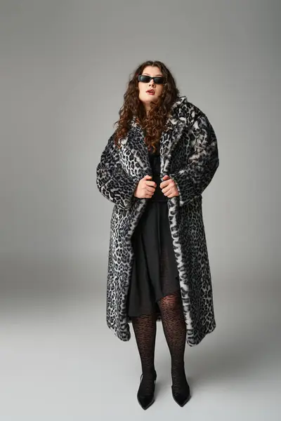 Hermosa mujer con curvas en abrigo de piel de leopardo y gafas de sol de pie sobre fondo gris - foto de stock