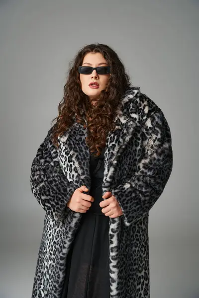 Attrayant plus la taille jeune femme en manteau de fourrure de léopard et lunettes de soleil debout sur fond gris — Photo de stock