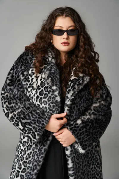 Élégant plus la taille jeune femme en manteau de fourrure de léopard et lunettes de soleil debout sur fond gris — Photo de stock