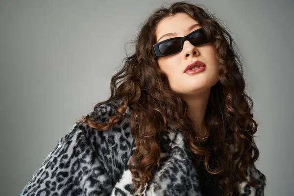 Ritratto di donna elegante plus size in pelliccia di leopardo e occhiali da sole appoggiati in avanti sul grigio — Foto stock