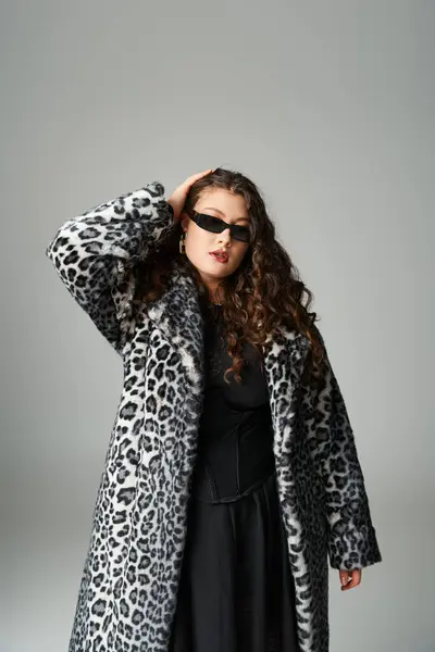 Hermosa mujer joven con curvas en abrigo de piel de leopardo y gafas de sol posando con la mano en la cabeza en gris - foto de stock