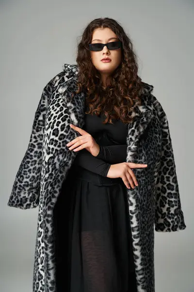 Mujer con curvas seguras en gafas de sol de pie con abrigo de piel de leopardo sobre fondo gris - foto de stock