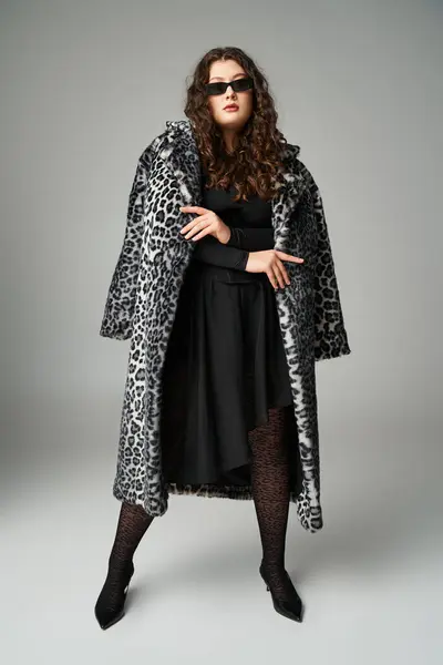 Belle femme plus la taille dans des lunettes de soleil debout avec manteau de fourrure de léopard sur fond gris — Photo de stock