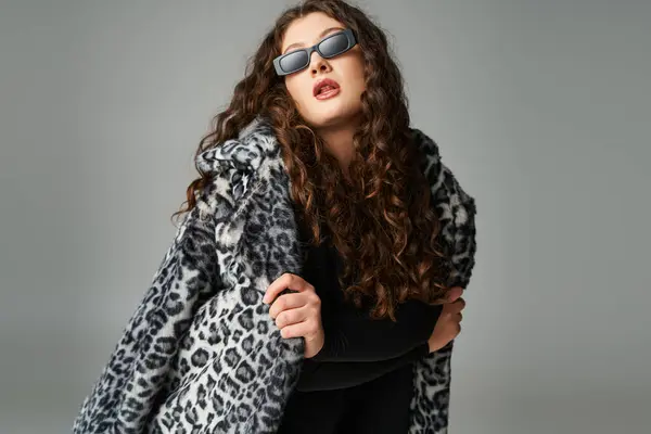 Lovely plus size woman in sunglasses leaning forward with leopard fur coat on grey background — Stock Photo