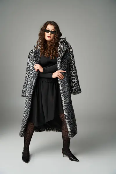 Mulher curvilínea em óculos de sol de pé com casaco de pele de leopardo e pernas afastadas no fundo cinza — Fotografia de Stock