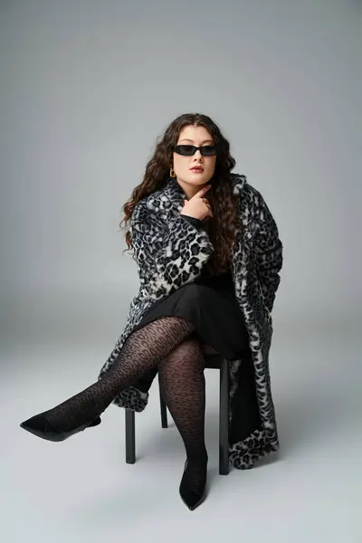 Confiant plus la taille jeune femme en manteau de fourrure de léopard et lunettes de soleil assis sur chaise noire — Photo de stock